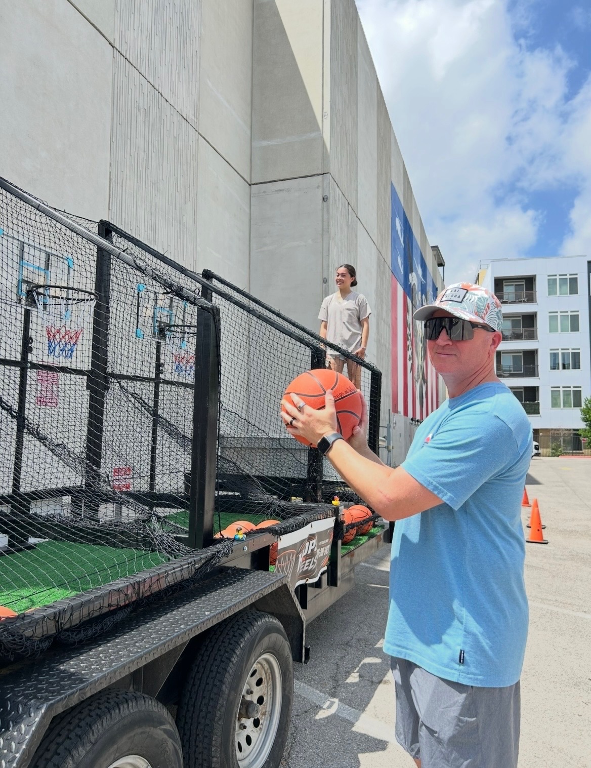 A guest holding a basketball beside the Hoops on Wheels trailer, ready to take a shot at one of the four stations