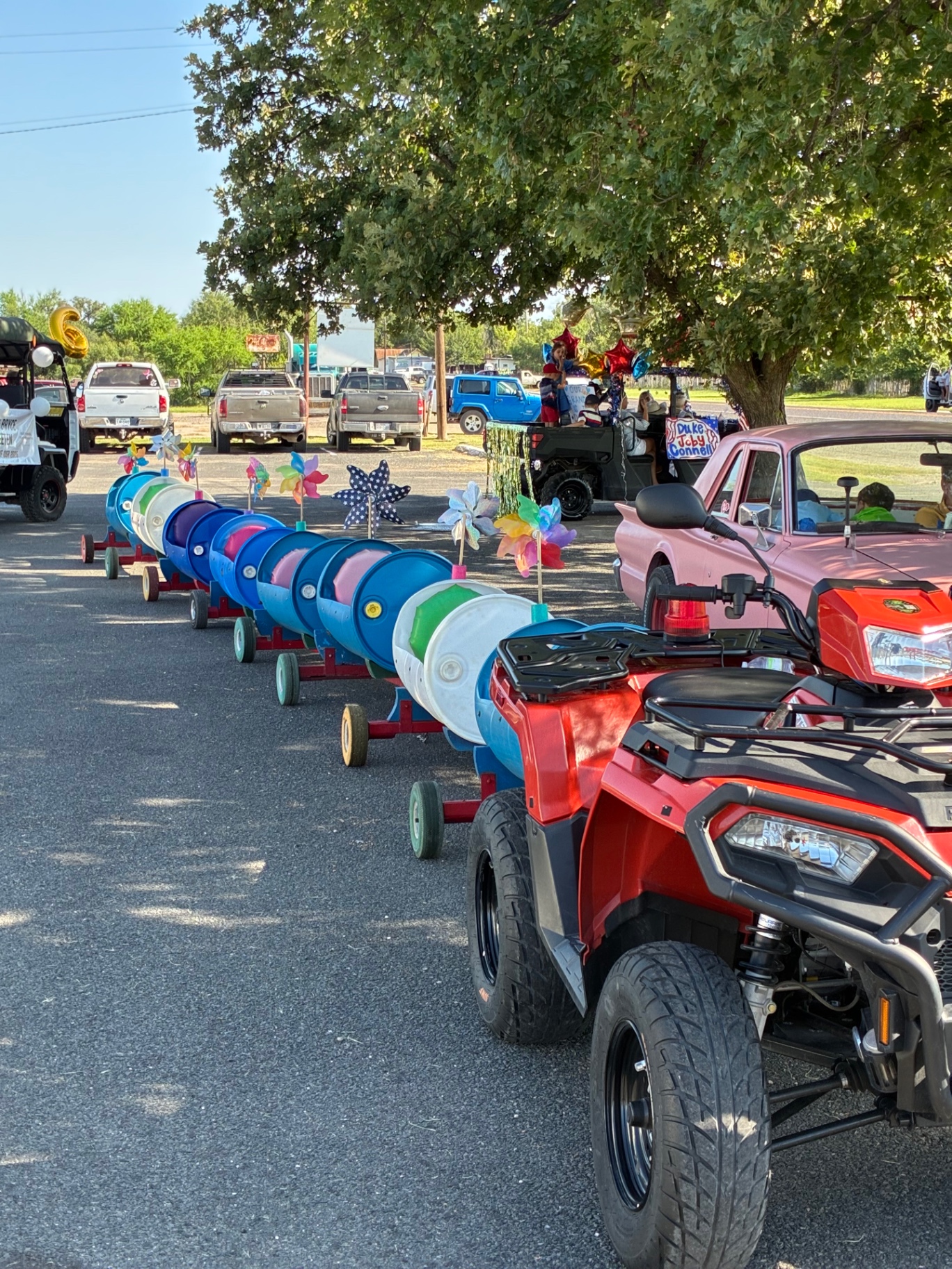 The Doodle Train cars lined up on pavement behind the red ATV, decorated with pinwheels, ready to roll at a community parade