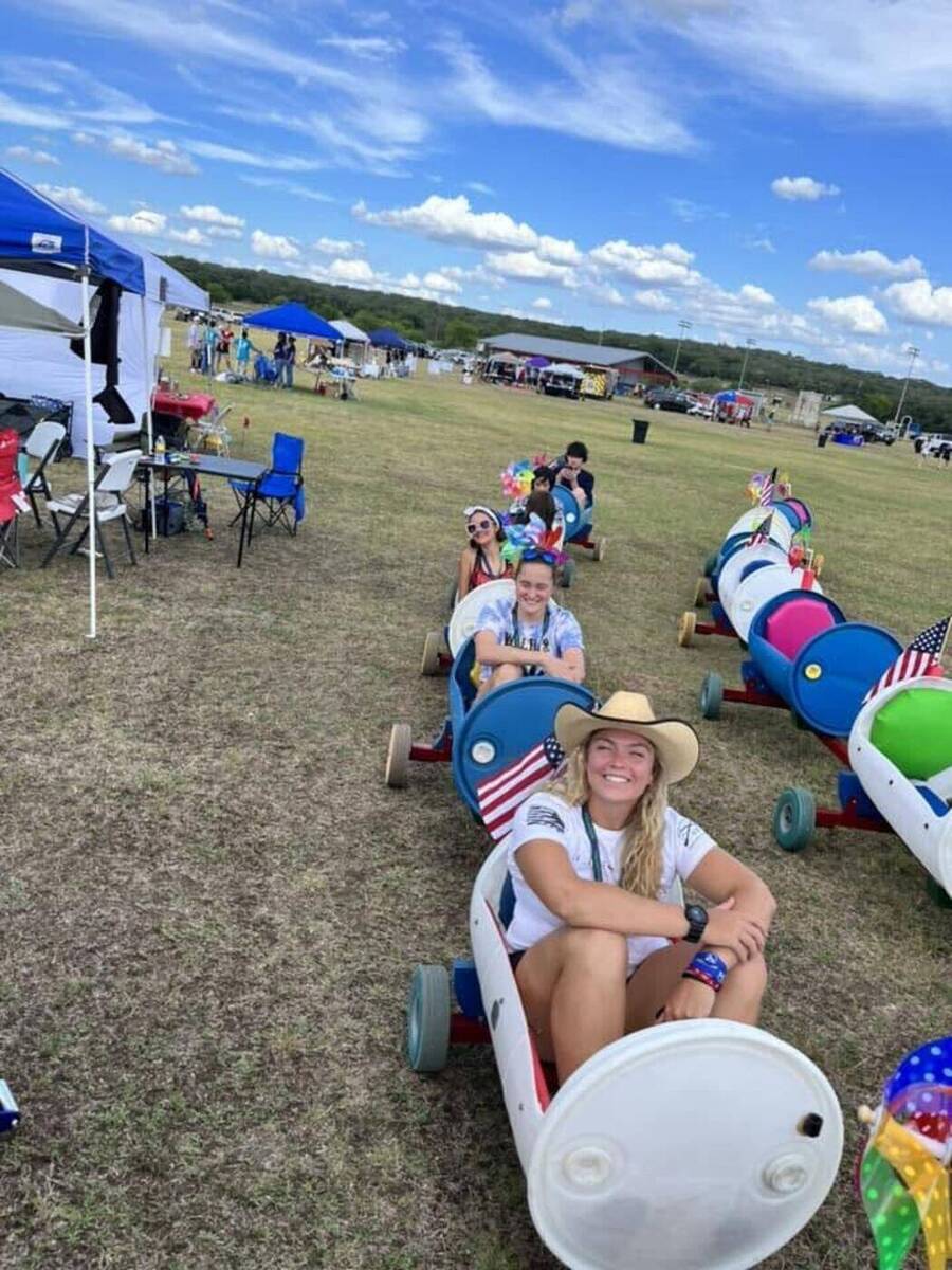 A line of smiling adults riding the colorful Doodle Train at an outdoor community event under a bright blue sky